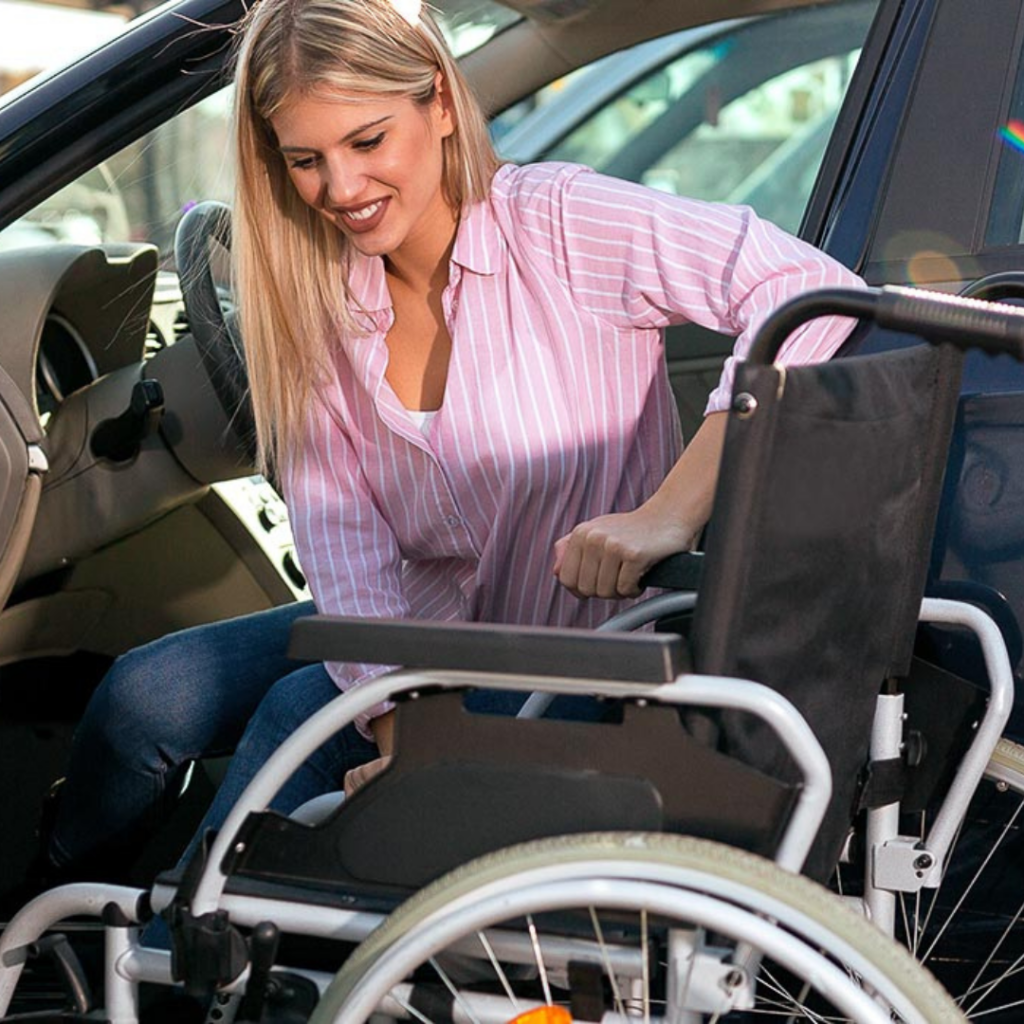 Eine blonde Frau sitzt auf der Fahrer-Seite von einem Auto. Neben ihr steht ein Rollstuhl. Sie stemmt sich auf der Arm-Lehne ab, damit sie sich in den Rollstuhl setzen kann.